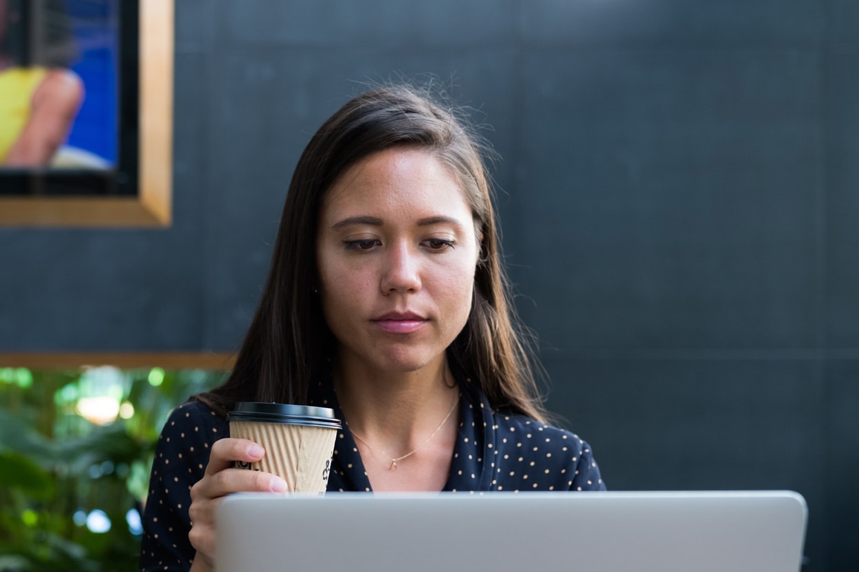 woman sitting at a cafe holding a coffee while using a laptop computer